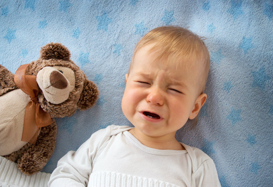 Upset toddler lying in bed with teddy bear grimacing